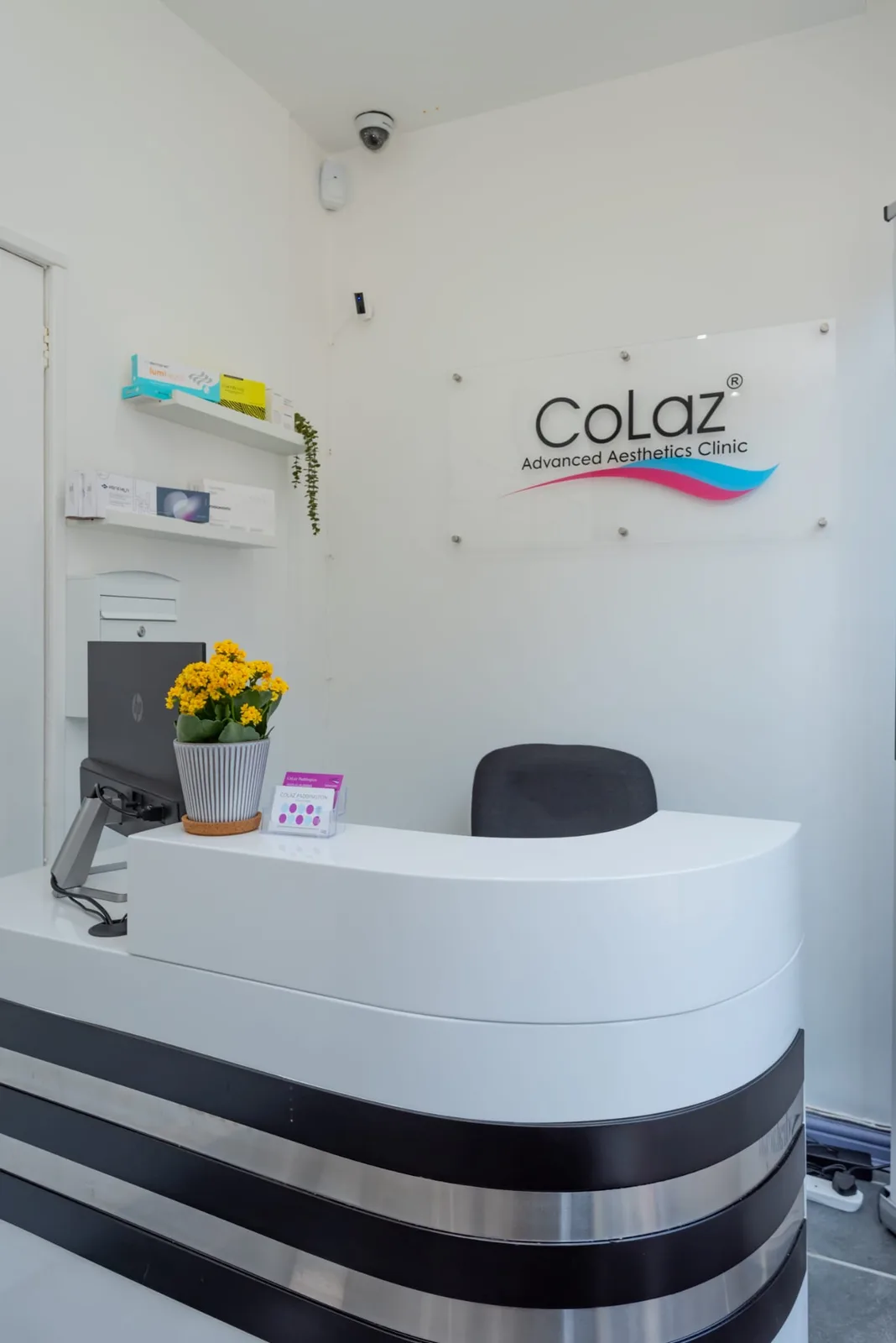 Reception at the CoLaz London Paddington clinic, with the curved white desk, a pot of yellow flowers and the CoLaz Advanced Aesthetics Clinic sign on the wall behind