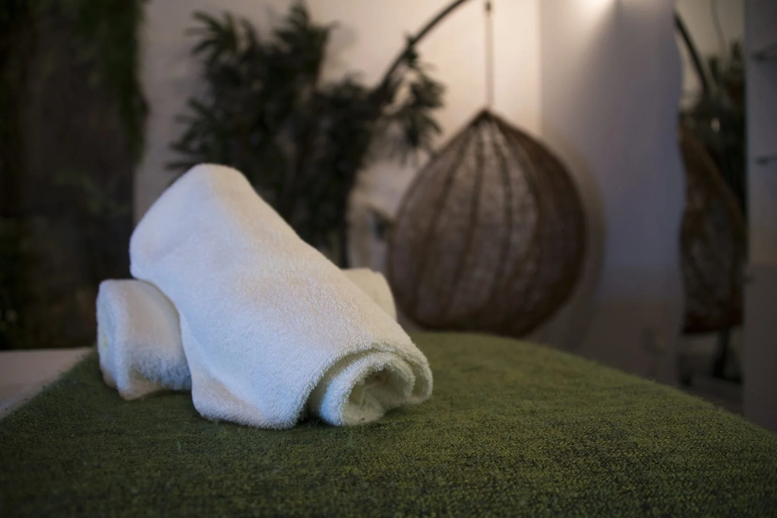 Detail of fresh white towels on a green textured bench in the Southall clinic relaxation lounge, with rattan hanging chairs and plants in the background