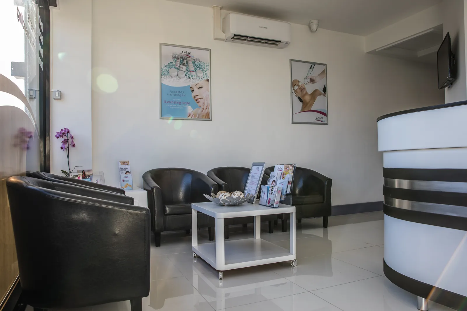 Reception and waiting area inside the CoLaz Wembley clinic, with black leather tub chairs, a white coffee table and treatment posters on the wall beside the reception desk